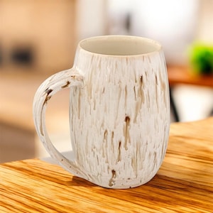 May include: A cream-coloured ceramic mug with a large handle. The mug features brown drips and speckles. The mug is on a wooden surface.
