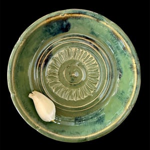 May include: A handmade, green ceramic garlic grater with a central textured design and a small garlic clove. The grater has a circular shape with a slightly raised rim and a glossy finish. The color is a mix of green and blue.