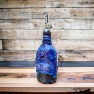 May include: A ceramic oil bottle with a stainless steel spout. The bottle has a striking blue glaze with purple and black details. The design includes a textured neck and a dark base, suitable for kitchen use.