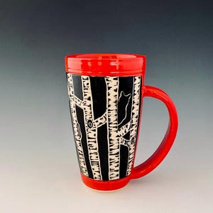 May include: A ceramic travel mug with a red handle, rim, and base. The mug features a black and white birch tree design with a small fox silhouette. The mug is designed for hot or cold beverages.