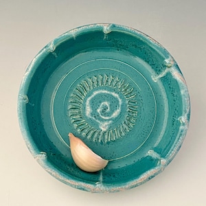 May include: A teal ceramic garlic grater with a spiral grating surface and a raised rim. A single garlic clove rests on the grater. The grater is handmade and has a rustic, artisanal appearance.