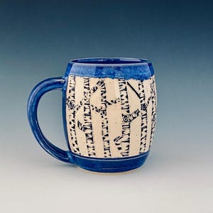 May include: A ceramic mug with a blue handle and rim. The mug has a cream-coloured body with a black tree design. The mug is approximately 10 cm tall and 9 cm in diameter, suitable for hot beverages.