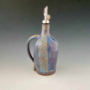 Op de afbeelding: Een handgemaakte keramische oliefles met een blauwe en paarse glazuur. De fles heeft een decoratief patroon en een zilveren tuit. Het heeft een handvat voor gemakkelijk schenken en is een stijlvol keukenaccessoire.