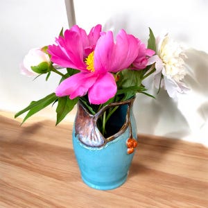 May include: A turquoise ceramic vase with a brown leafy design and orange berries. The vase is filled with pink and white peonies.