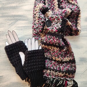 May include: A handmade crochet scarf and matching fingerless gloves set. The scarf is a mix of black, pink, beige, and green yarn with a button closure. The fingerless gloves are solid black. The scarf has a fringe at the bottom.
