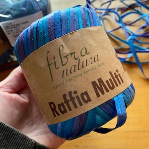 May include: A skein of raffia yarn in shades of blue and purple, wrapped with a brown paper label. The label reads "fibra natura Raffia Multi" and "Natural Fine Hand Knitting Yarns".