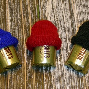 Pu&ograve; includere: Tre barrette di cioccolato Hershey's, ciascuna avvolta in carta dorata e sormontata da un mini cappello lavorato a maglia. I cappelli sono blu, rossi e neri. Le barrette sono disposte su una superficie di legno rustico color chiaro.