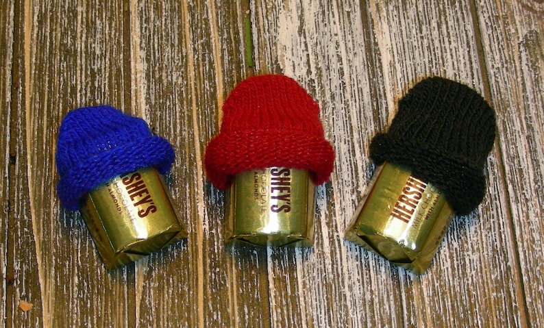 Pu&ograve; includere: Tre barrette di cioccolato Hershey's, ciascuna avvolta in carta dorata e sormontata da un mini cappello lavorato a maglia. I cappelli sono blu, rossi e neri. Le barrette sono disposte su una superficie di legno rustico color chiaro.