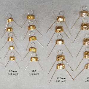 May include: A collection of gold-colored ornament caps with silver wire hangers. Sizes range from 6.5mm (.25 inch) to 15 mm (.59 inch). The caps have a decorative pattern and are designed for Christmas ornaments.