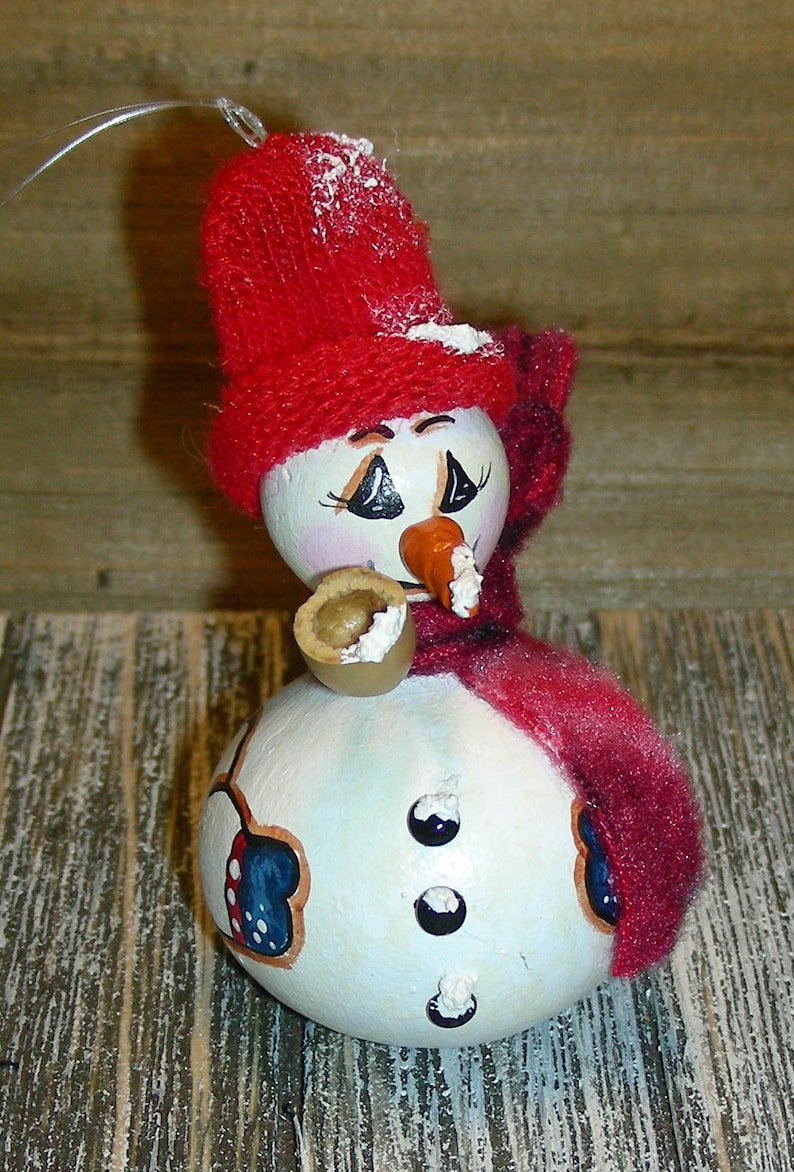Pu&ograve; includere: Ornamento a forma di pupazzo di neve dipinto a mano con cappello e sciarpa rossi a maglia. Il pupazzo di neve ha un naso a carota, occhi neri e una tazza piccola. L'ornamento &egrave; decorato con dettagli blu e rossi e tre bottoni neri.