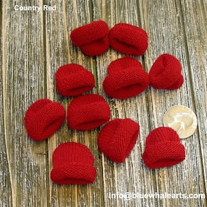 Pu&ograve; includere: Una collezione di piccoli cappelli rossi lavorati a maglia, etichettati come "Country Red", sono sparsi su una superficie di legno. I cappelli sono in miniatura, con un bordo ripiegato. Una moneta &egrave; inclusa come riferimento delle dimensioni. L'indirizzo email info@bluewhalearts.com &egrave; visibile.