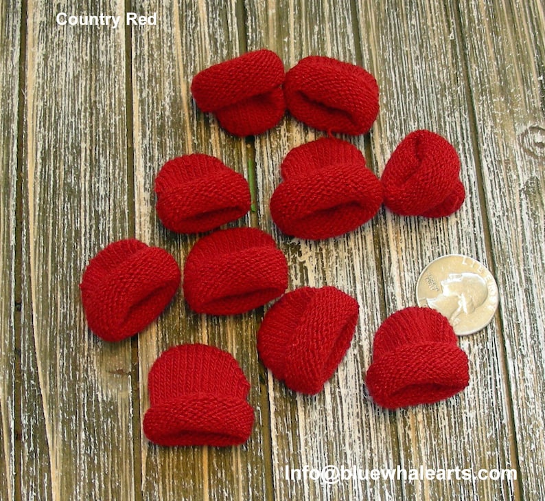 Pu&ograve; includere: Una collezione di piccoli cappelli rossi lavorati a maglia, etichettati come "Country Red", sono sparsi su una superficie di legno. I cappelli sono in miniatura, con un bordo ripiegato. Una moneta &egrave; inclusa come riferimento delle dimensioni. L'indirizzo email info@bluewhalearts.com &egrave; visibile.