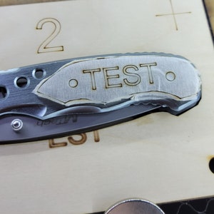 May include: A silver pocket knife with a black handle and a laser engraved inscription that reads "TEST".