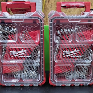 Puede incluir: Dos organizadores de herramientas Milwaukee Packout rojos y transparentes. Cada estuche tiene un asa roja, una tapa transparente y múltiples compartimentos llenos de brocas plateadas. El logotipo de Milwaukee y "PACKOUT" están impresos en blanco.