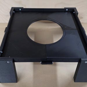 Könnte beinhalten: Ein schwarzer, rechteckiger Rahmen mit einer großen kreisförmigen Öffnung in der Mitte. Der Rahmen wird von vier rechteckigen Beinen getragen, eines an jeder Ecke. Der Rahmen ist mit Schrauben konstruiert und scheint aus einem Verbundwerkstoff zu bestehen.