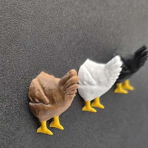 May include: Three chicken figurines in a row, each with yellow feet. The first is brown, the second is white, and the third is black. The figurines are likely magnets or decorative items.