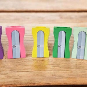 Könnte beinhalten: Sechs bunte Anspitzer in Lila, Rosa, Gelb, Grün, Hellgrün und Türkis. Jeder Anspitzer hat eine silberne Klinge und eine Schraube. Sie sind auf einer Holzoberfläche angeordnet.