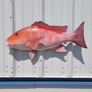 May include: A vibrant, reddish-pink fish sculpture with detailed fins and scales. The fish is mounted on a white corrugated metal surface with a blue accent. The sculpture's open mouth and prominent fins add to its realistic appearance.