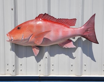 May include: A vibrant, reddish-pink fish sculpture with detailed fins and scales. The fish is mounted on a white corrugated metal surface with a blue accent. The sculpture's open mouth and prominent fins add to its realistic appearance.