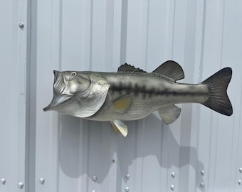 May include: A wall-mounted fish sculpture featuring a largemouth bass with its mouth wide open. The fish is painted in shades of silver, gray, and dark green with a prominent horizontal stripe along its side and detailed fins.
