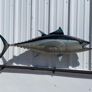 May include: A large, blue and silver, metal fish sculpture mounted on a white wall. The fish has a long, slender body with a pointed head and a large, open mouth.