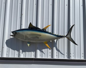 May include: A realistic, painted sculpture of a tuna fish, mounted on a white corrugated metal wall. The fish features a blue and silver body with yellow and black fins. The mouth is open, and the tail is extended.