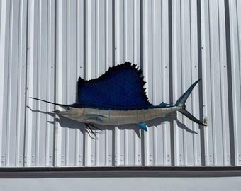 May include: A blue and silver sailfish mounted on a white corrugated metal wall. The sailfish has a long, thin bill and a large, pointed dorsal fin.