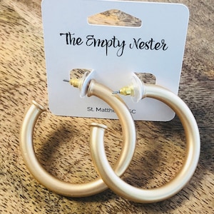 May include: A pair of gold hoop earrings with a matte finish. The earrings are on a white card with the text "The Empty Nester" printed on it.