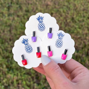 May include: Six miniature nail polish bottle earrings on white cards with pineapple designs. The earrings are in shades of pink, red, and purple.