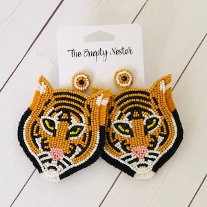 May include: A pair of beaded tiger head earrings with orange, black, white, and yellow beads. The earrings have a stud post and a matching beaded stud. The tag reads "The Empty Nester".