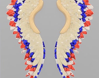 May include: A pair of gold-toned earrings with red, white, and blue glitter wings. The wings are shaped like angel wings and have a layered look.