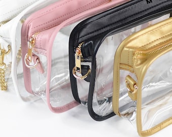 Peut inclure: Quatre sacs en plastique transparent avec des bordures en cuir de différentes couleurs. Les sacs ont des fermetures éclair et des chaînes dorées. Les sacs sont alignés de gauche à droite : blanc, rose, noir et or.