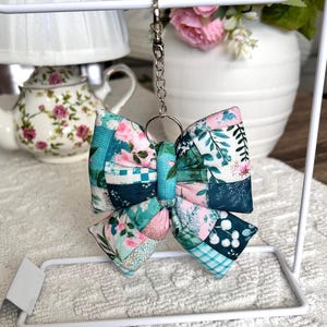 May include: A handmade bow-shaped keychain with a patchwork design. The bow features a mix of floral and geometric patterns in shades of teal, pink, and white. It hangs from a silver-colored chain and ring. The background includes a white stand and floral arrangement.