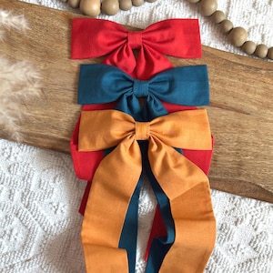 May include: Three fabric hair bows in red, teal, and orange. The bows are arranged on a wooden surface, with the orange bow having long, flowing tails. The bows are made of a soft fabric and are likely handmade.