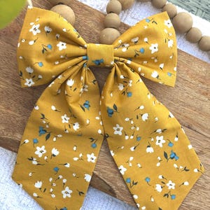 May include: Mustard yellow fabric bow with a floral pattern of white and blue flowers. The bow is tied in the center and has long, pointed tails. The fabric has a slight sheen, and the bow is likely handmade.