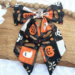 May include: A large Halloween-themed bow tie featuring a patchwork design with pumpkins, ghosts, and skulls on a black background. The bow tie has orange, white, and brown accents, perfect for festive occasions.