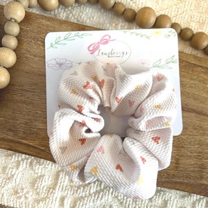 May include: A white scrunchie with a small heart pattern in shades of pink, orange, and yellow. The scrunchie is made of a soft, textured fabric. The scrunchie is on a white card with the text "Lansberry's Boutique" in pink.