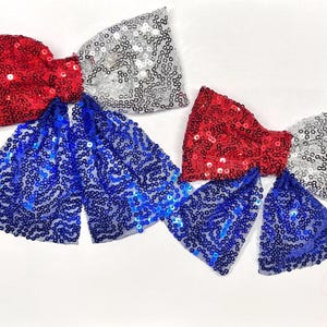 May include: Several sequined bow ties in red, silver, and blue are displayed against a white background. The bows are arranged in a patriotic color scheme, with each bow featuring a different color combination. The sequins add a sparkling, festive touch.