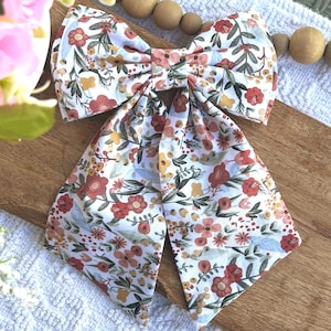 May include: A large, floral patterned bow tie featuring a white background with red, pink, and yellow flowers and green leaves. The bow tie is made of fabric and has long, flowing tails. The bow tie is a hair accessory.