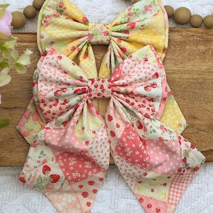 May include: Two fabric hair bows featuring a patchwork design with strawberry patterns, small floral prints, and gingham checks in shades of yellow, pink, and white. The bows have long, pointed tails and are constructed from multiple cotton fabric segments.