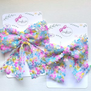 May include: Two large, colorful hair bows with iridescent sequins. The bows are attached to white cards with the text "Leasberys Boutique" and floral accents. The bows feature a variety of pastel colors, including pink, blue, yellow, and green.