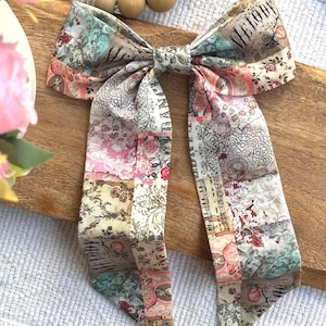 May include: A fabric hair bow featuring a collage of vintage-inspired patterns including floral motifs, musical notes, script text, and the word MELODY. The bow has two long, dangling tails and is constructed from a multi-colored textile with muted tones.