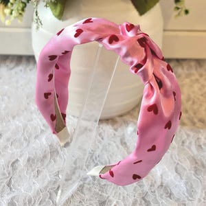 May include: A pink headband with a knotted top, adorned with small red hearts. The headband is made of a silky material and has white tips on the ends. The accessory is displayed on a clear stand.