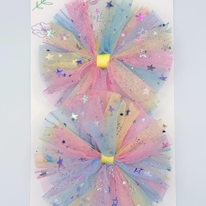 May include: Two round hair accessories made of layered, multicolored tulle fabric in shades of pink, blue, and yellow. The tulle is decorated with small, holographic star-shaped sequins and glitter. Each piece features a yellow ribbon center.