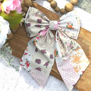 May include: A large fabric hair bow featuring a patchwork design of various floral patterns in shades of pink, orange, and green, interspersed with white polka dot sections on a cream background, finished with a central fabric knot detail.