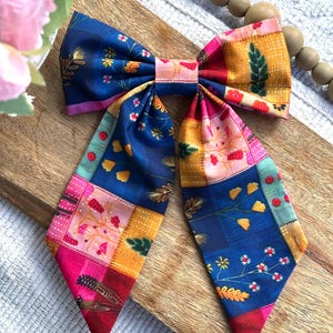 May include: A fabric hair bow featuring a patchwork design with various floral and botanical patterns in shades of deep blue, mustard yellow, bright pink, and red. The bow has two long, pointed tails and a central knot detail made of the same fabric.