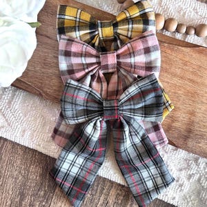 May include: Three plaid fabric bows in yellow, pink, and gray, arranged on a wooden surface. The bows have a classic bow tie shape with a plaid pattern in various colors. The background includes a white fabric and wooden beads.