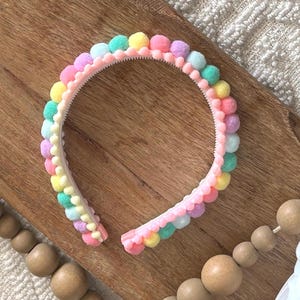 May include: A pastel-colored headband with a row of small pom-poms in shades of pink, purple, yellow, and teal. The headband is made of a light pink plastic and is designed for wear. The pom-poms are evenly spaced along the top of the headband.