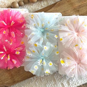 May include: Three sets of hair bows in pink, light blue, and pale pink. Each bow is made of layered tulle with a daisy pattern. The bows are attached to white cards, perfect for gifting or display.