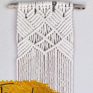 May include: White macrame wall hanging with a geometric pattern. The macrame is hung on a wooden dowel rod.
